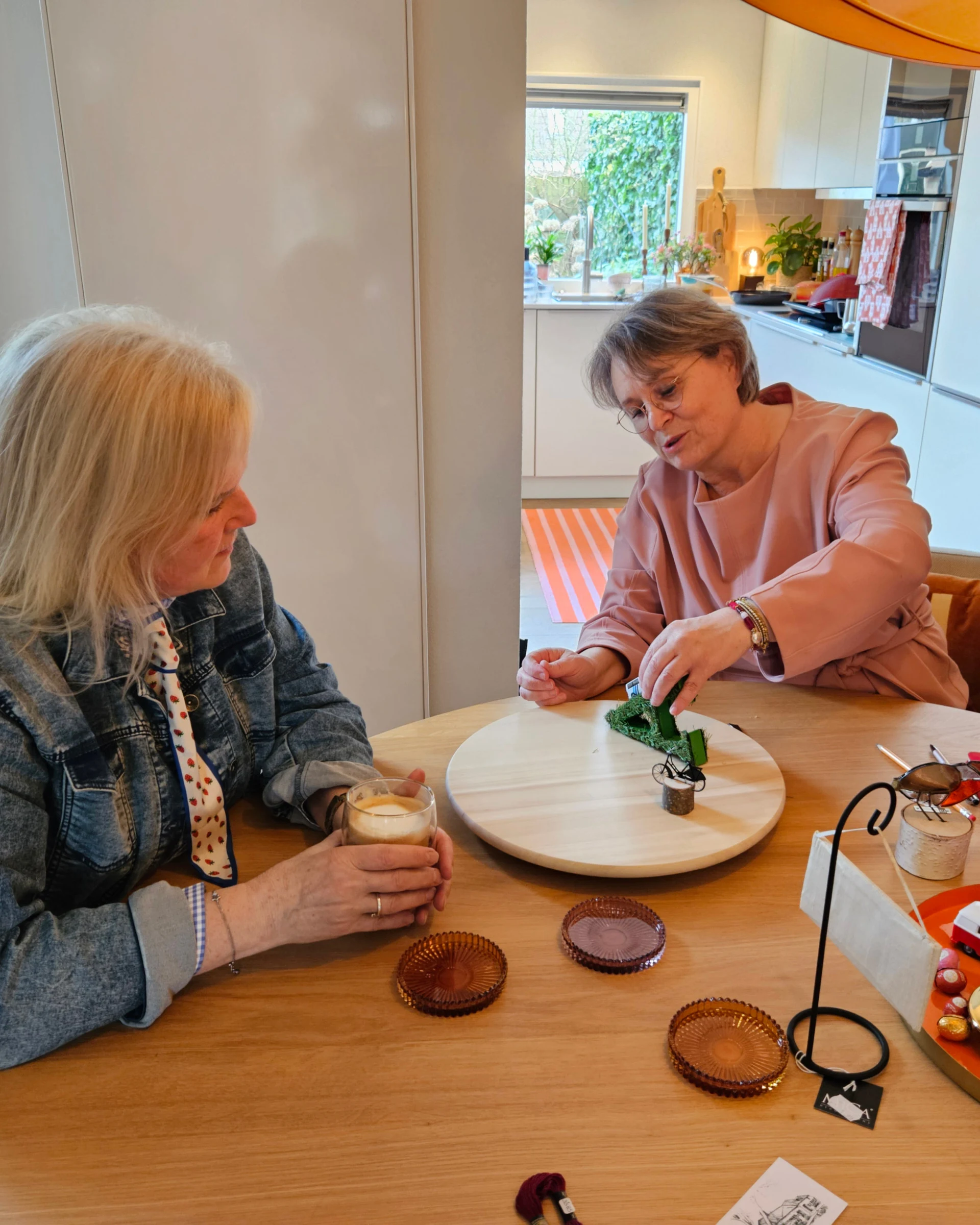 Twee vrouwen aan een ronde tafel; de één werkt aan een opstelling op een draaiplateau, de ander kijkt met een kopje koffie toe.