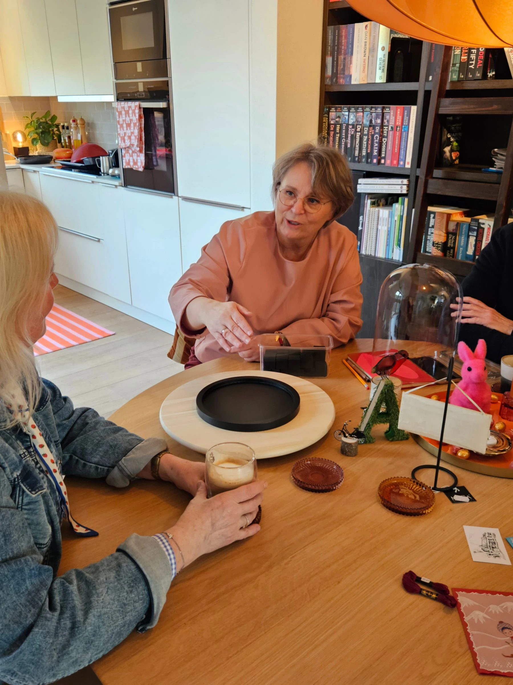 Drie mensen in gesprek aan een ronde tafel, met een glazen stolp en een paar voorwerpen tussen hen in.