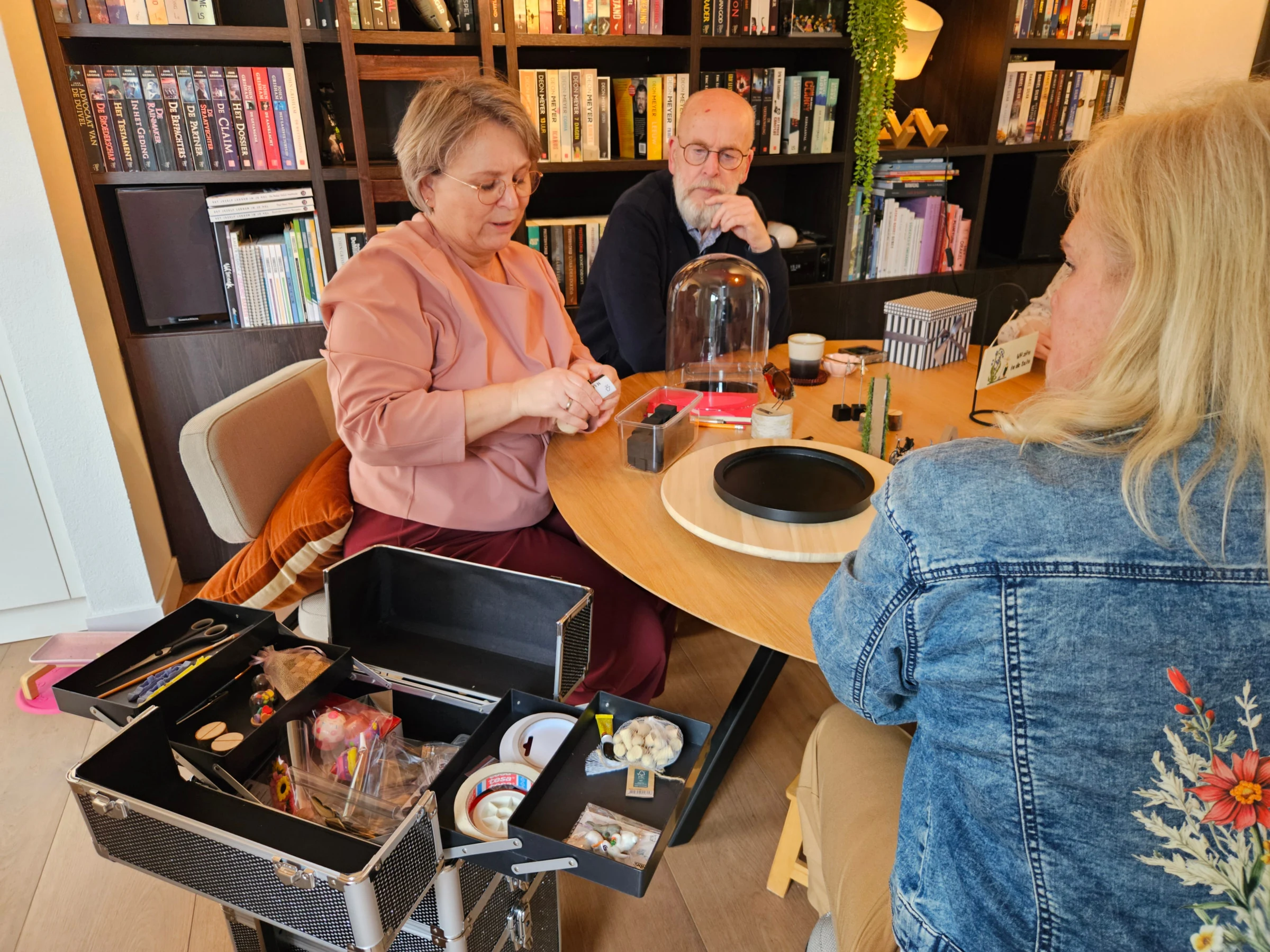Aan een ronde tafel voor een boekenkast wordt gewerkt aan een stolp; naast de tafel staat een open koffer vol materialen.
