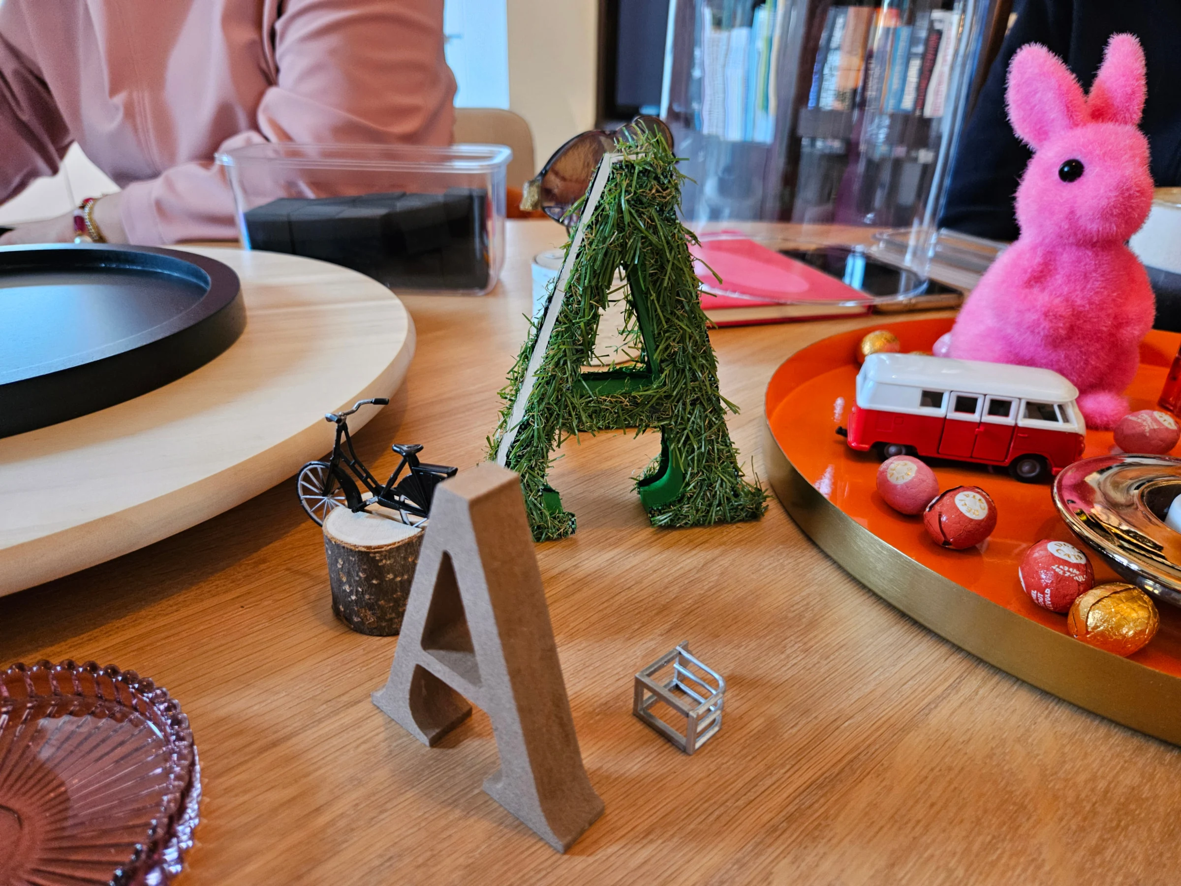 Voorwerpen op tafel: een letter A van mos met een fietsje, een houten letter A, een rood-wit speelgoedbusje en een roze haasje.