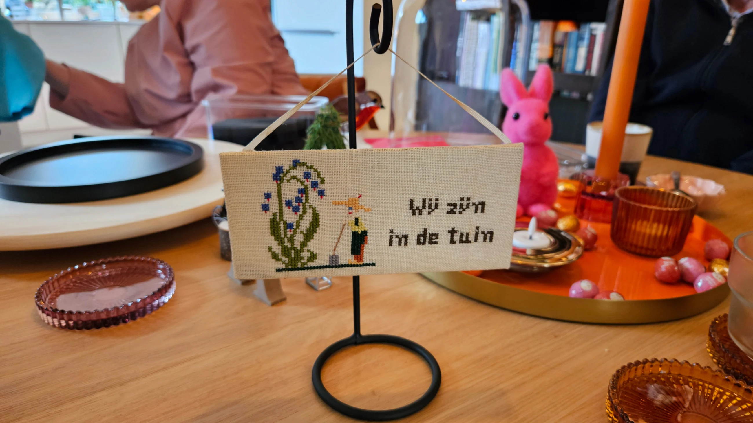 Een geborduurd lapje met de tekst 'Wij zijn in de tuin' aan een zwart standaardje, met daarachter een roze haasje, een kaars en een glazen stolp.